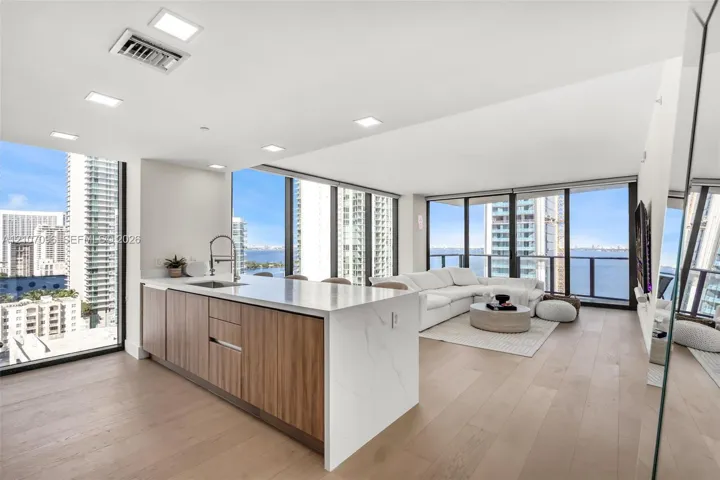 Kitchen featuring a center island with light-toned stone-finish countertop, wood-finish cabinetry, under-mount sink, and recessed lighting throughout, complemented by wood-finish flooring and expansive floor-to-ceiling windows