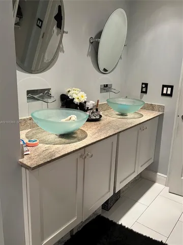 Dual vanity featuring white shaker-style cabinetry, a stone countertop, wall-mounted faucets, frosted glass vessel sinks, and oval mirrors
