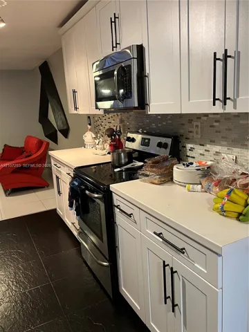 White shaker-style cabinetry with matte black pulls, solid surface countertops, a mosaic tile backsplash, and stainless steel kitchen appliances