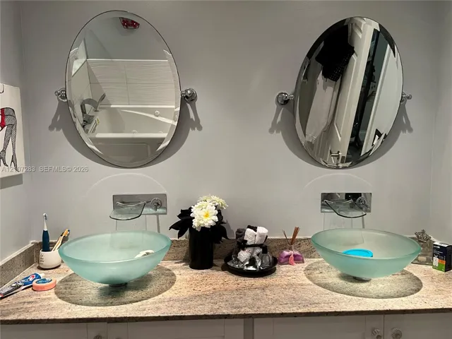 Dual vanity featuring frosted glass vessel sinks, stone countertop, wall-mounted faucets, and oval tilting mirrors