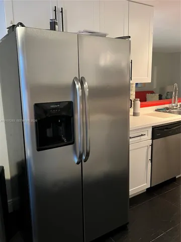 Stainless steel side-by-side refrigerator, white shaker-style cabinetry with matte black hardware, stone-finish countertops, undermount sink, and dark tile flooring