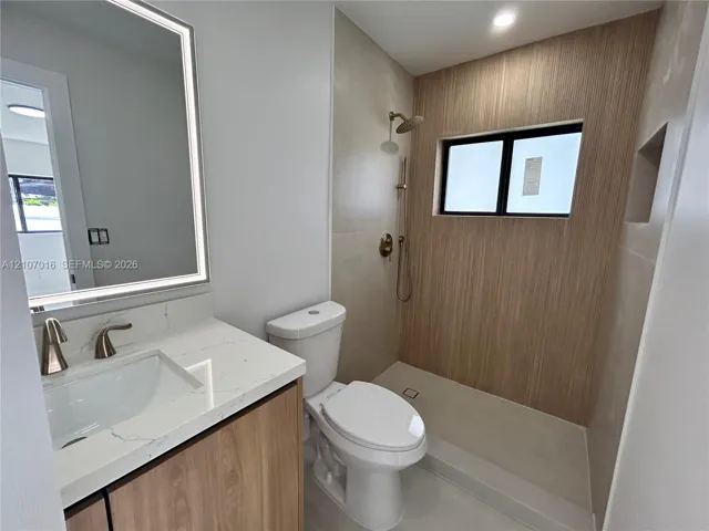 Bathroom featuring a vanity with a white countertop and gold-toned faucet, a wall-mounted LED mirror, and a walk-in shower with vertical wood-finish wall tile, gold-toned fixtures, and niche.