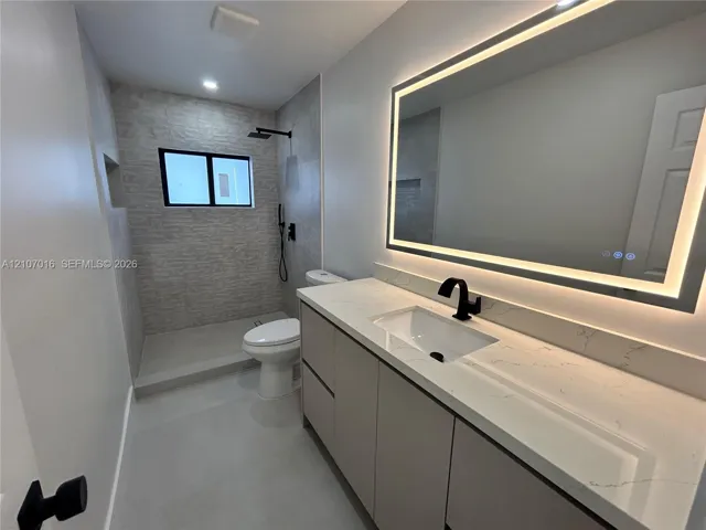 Modern vanity featuring a stone-composite countertop with integrated sink, sleek cabinetry, a large LED-backlit mirror, and matte black fixtures