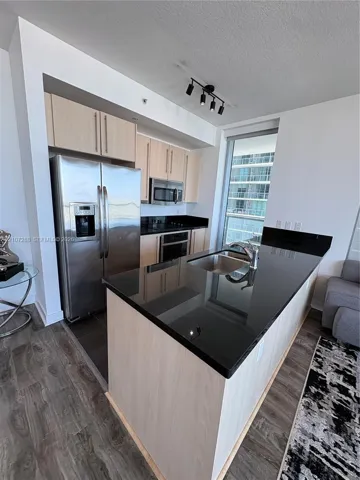 Kitchen featuring light wood-finish cabinetry, black stone countertops, stainless steel appliances, a center island with an undermount sink, and track lighting