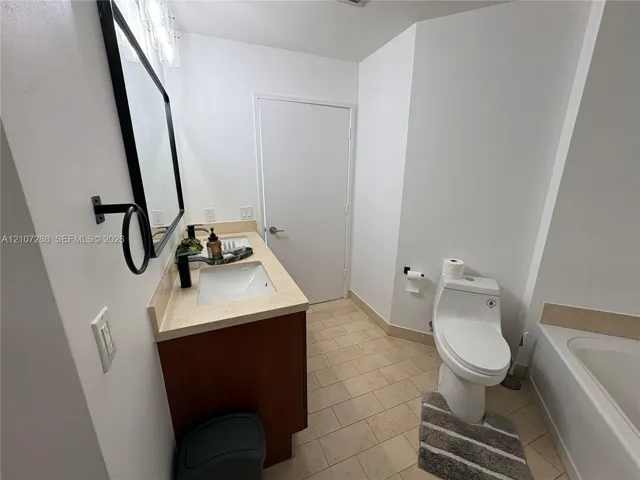 Bathroom featuring a wood-finish vanity with a stone-composite countertop, an integrated rectangular sink, a black-framed mirror, and ceramic tile flooring