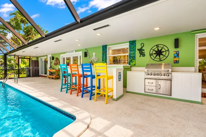 Screened lanai featuring a built-in outdoor kitchen with stainless steel grill, composite bar-height counter, recessed ceiling lighting, and pool with aggregate decking