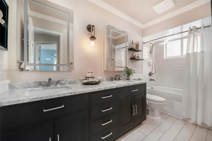 Hallway Bath. Double vanity featuring dark cabinetry, stone-finish countertops, lighted dual mirrored cabinets, wall-mounted lighting, and a bathtub-shower combination.