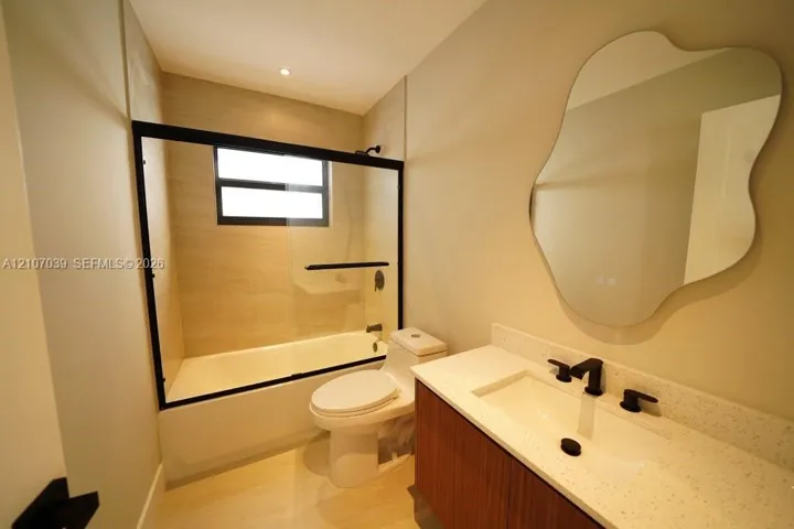 Floating vanity with light-colored speckled countertop, undermount sink, matte black widespread faucet, organic-shaped wall mirror, and tub-shower combination featuring a black-framed glass enclosure