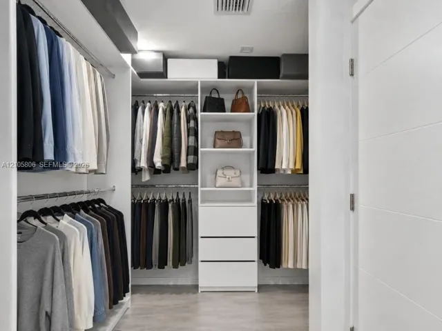 Custom walk-in closet featuring white built-in shelving, a central cabinetry unit with three drawers, dual-tier metal hanging rods, and wood-finish flooring