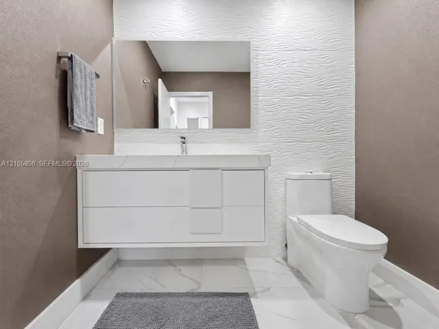 Floating vanity with integrated sink, stone-surface countertop, textured white feature wall, large-format tiled flooring, and dual-flush toilet