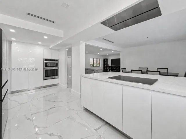 Modern kitchen featuring a central island with an integrated cooktop, stone-finish countertops, high-gloss cabinetry, stainless steel double wall ovens, and large-format tile flooring