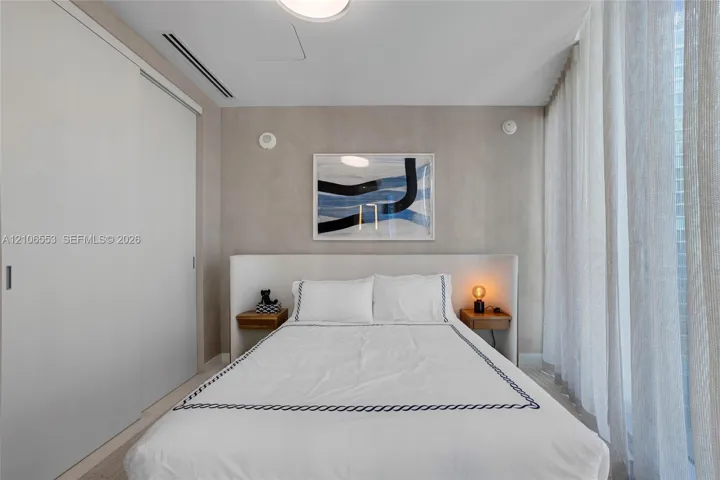 Neutral-toned room featuring a built-in upholstered headboard, dual integrated wood-finish bedside shelves, floor-to-ceiling sliding closet doors, and light-filtering floor-to-ceiling drapery