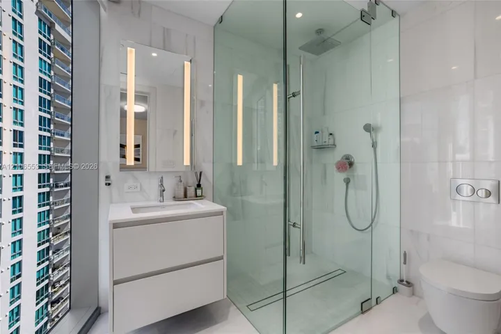 Bathroom featuring a floating vanity with a rectangular sink, wall-mounted mirror with integrated lighting, glass-enclosed shower with rainfall head and handheld sprayer, wall-mounted toilet, and floor-to-ceiling tile finishes
