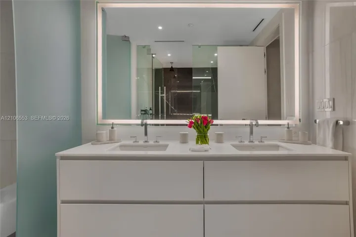 Double vanity featuring a white cabinetry base, dual undermount sinks, stone countertops, wall-mounted chrome faucets, and an expansive integrated LED-backlit mirror