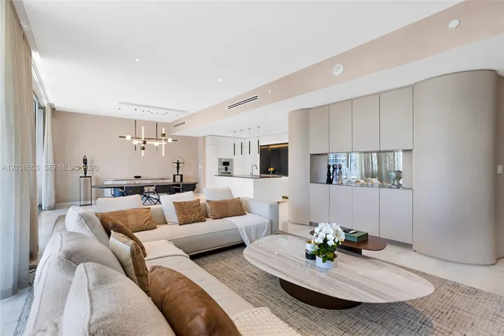 Open-concept living space featuring floor-to-ceiling windows, light-toned stone-finish flooring, custom curved built-in cabinetry, recessed lighting, and a modern multi-light chandelier
