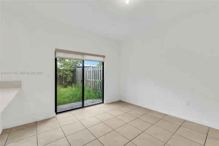 Interior featuring ceramic tile flooring, neutral walls, and a sliding glass door with a roller shade, leading to a fenced outdoor area with paver hardscaping
