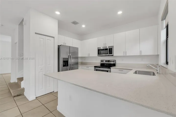 Kitchen featuring white flat-panel cabinetry, light-toned stone-finish countertops, stainless steel appliances, a peninsula with an integrated sink, and tile flooring