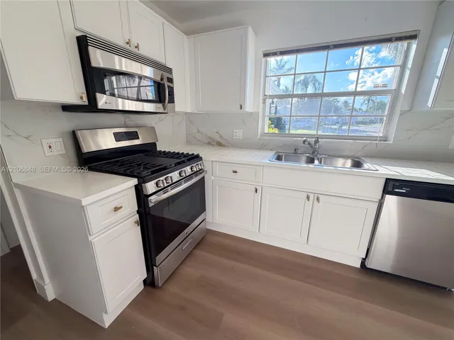 Kitchen featuring white shaker-style cabinetry, stone-patterned backsplash, stainless steel appliances, dual-basin sink, and wood-finish flooring
