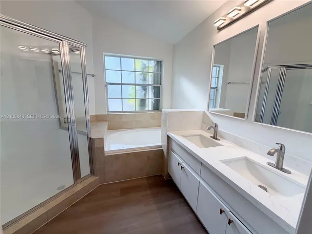 Double vanity with white quartz countertops, dual undermount sinks, wood-finish flooring, glass-enclosed shower, and built-in soaking tub with tile surround