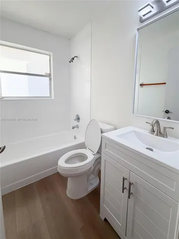 Bathroom featuring wood-finish flooring, white cabinetry with silver-tone hardware, a white-tiled shower surround, a rectangular wall-mounted mirror, and a single-basin sink with a metallic faucet