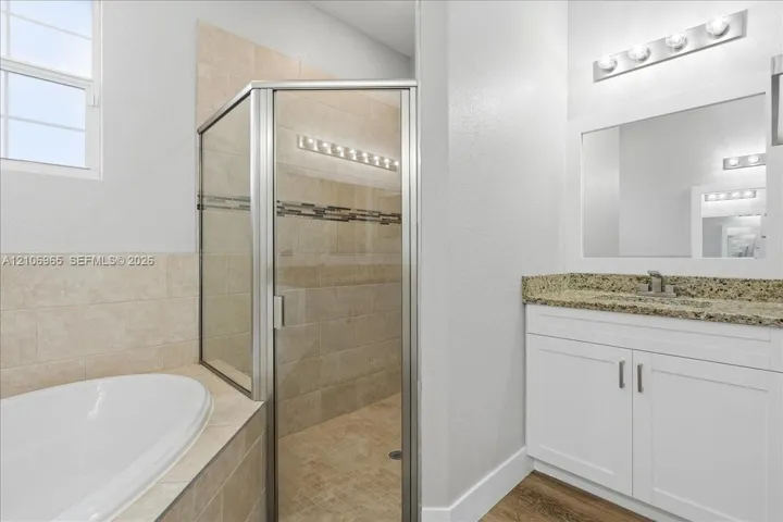Bathroom featuring a soaking tub, glass-enclosed shower with tiled surround, white shaker-style vanity, stone-finish countertop, and wood-finish flooring