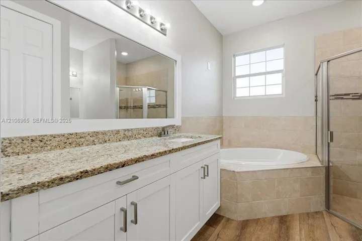 White vanity with a stone countertop, oversized wall-to-wall mirror, tiled soaking tub, walk-in glass-enclosed shower, and wood-finish flooring