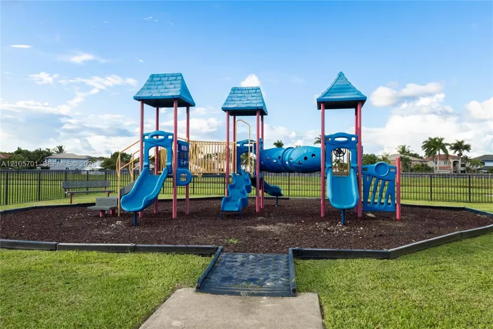 Multi-level playground structure featuring blue slides, a tunnel, and overhead canopies, situated on a contained mulch surface with metal perimeter fencing