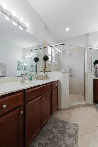Bathroom featuring dark wood-finish cabinetry, a light-toned countertop with an integrated sink, a full-width vanity mirror, tile flooring, and a glass-enclosed shower with wall-mounted hardware