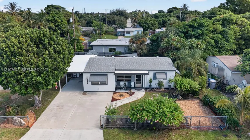 Single-story exterior with light-toned siding, concrete driveway, attached covered carport, metal window awnings, and fenced front yard