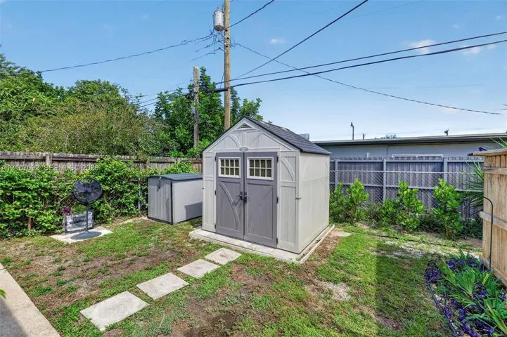 Exterior storage structure featuring a gable roof, double doors with glass paneling, and a reinforced foundation pad, situated within a fenced backyard