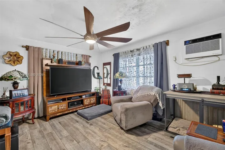 Living area featuring wood-finish flooring, a multi-blade ceiling fan with integrated lighting, and a wall-mounted air conditioning unit
