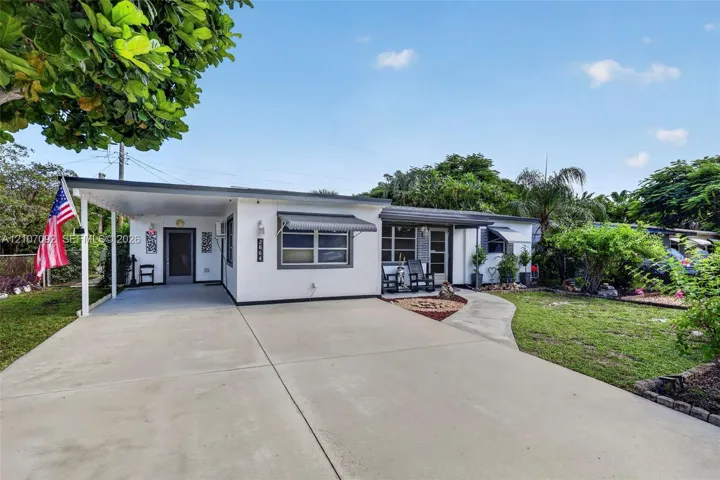 Single-story exterior featuring a concrete driveway, attached carport, metal window awnings, and a covered front entryway