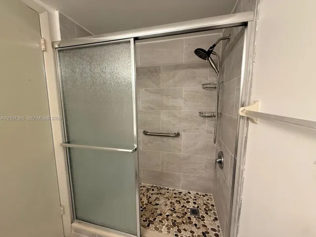 Walk-in shower featuring textured glass sliding doors, stone pebble floor tiling, large-format wall tile, a metal grab bar, and integrated storage shelving