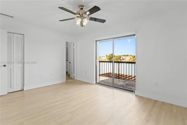 Interior space featuring light wood-finish flooring, a ceiling fan with integrated lighting, louvered-panel bi-fold doors, and a sliding glass door providing access to a railed balcony