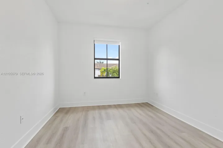 Neutral interior room featuring wood-finish flooring, crisp white walls, and a single window with black grid framing