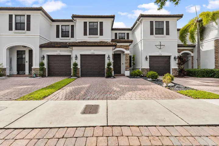 Mediterranean-inspired townhome exterior featuring multi-toned stone masonry accents, paver driveways, decorative shutters, and tile roofing
