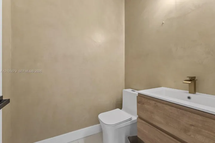 Textured plaster walls paired with a modern wood-finish vanity, integrated rectangular sink, brass-tone faucet, and a one-piece toilet