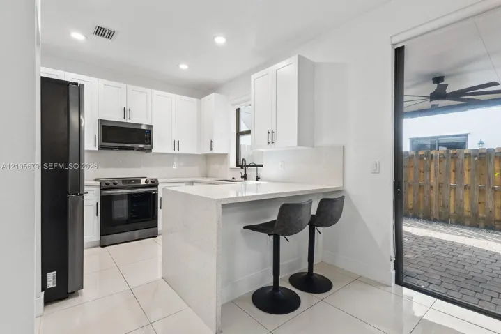 Kitchen featuring white shaker-style cabinetry, quartz countertops with an integrated peninsula, black stainless steel appliances, and large-format tile flooring with access to a paver patio