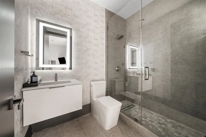 Floating vanity with a smooth-finish countertop, integrated rectangular sink, wall-mounted illuminated mirror, frameless glass-enclosed shower featuring textured tile work, and hexagonal floor tile