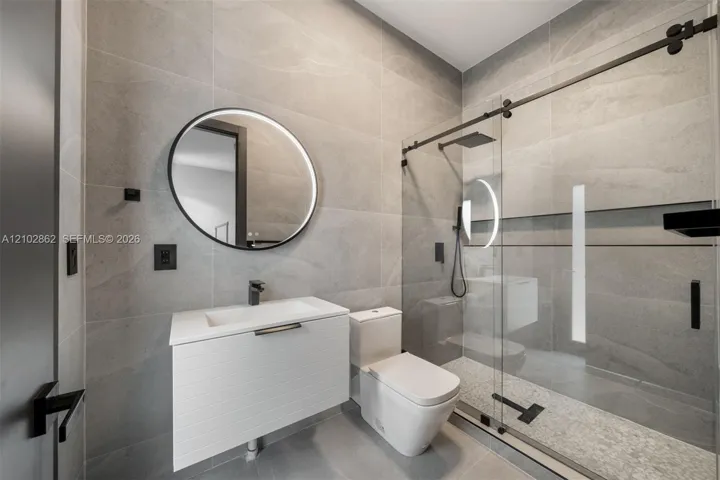 Floating white vanity with integrated basin, floor-to-ceiling large-format stone-finish tile, round backlit mirror, and glass-enclosed shower with matte black hardware