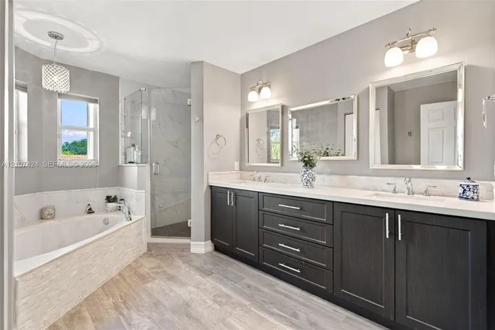 Dual-sink vanity with dark cabinetry, light-toned countertop, and framed mirrors, featuring a separate soaking tub with stone-textured surround and a glass-enclosed shower with tiled interior