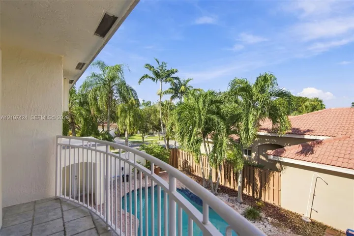 Covered balcony featuring stone-tile flooring and a white metal railing overlooking a private pool, wood fencing, and mature palm trees