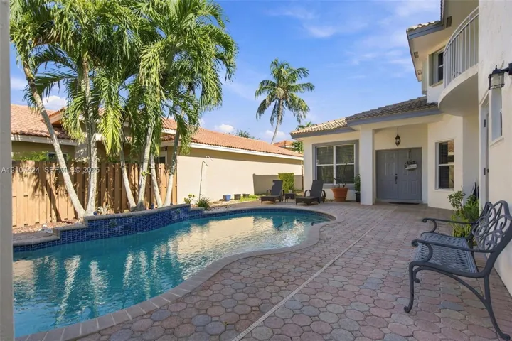 Rear exterior featuring a freeform swimming pool with blue tile accents, expansive paver patio, wood privacy fencing, and mature palm trees