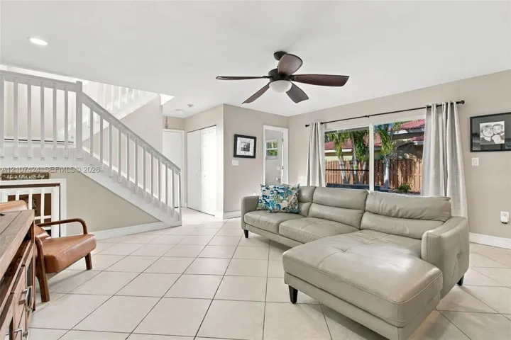 Living area featuring large-format ceramic tile flooring, a white wooden staircase with balustrades, a ceiling fan with integrated lighting, and multiple windows