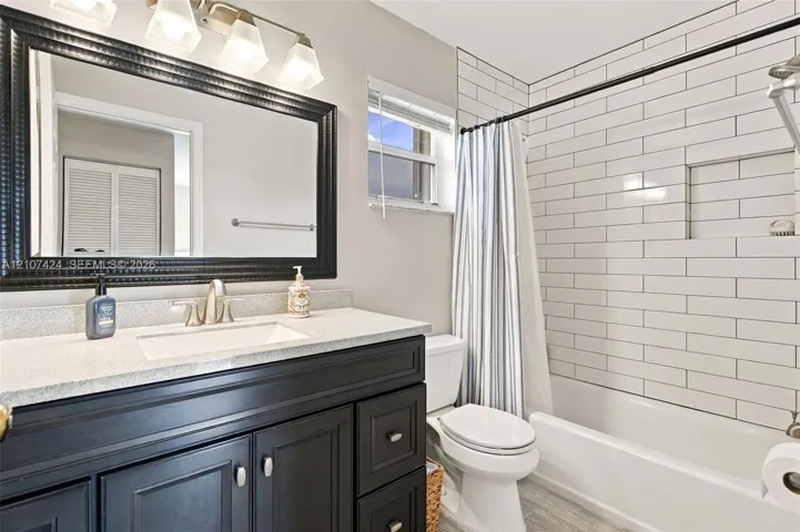 Bathroom featuring a dark-toned vanity with a light stone-surface countertop, rectangular undermount sink, framed wall mirror with integrated multi-bulb lighting, and a shower-tub combination finished with horizontal subway tiling and a recessed niche
