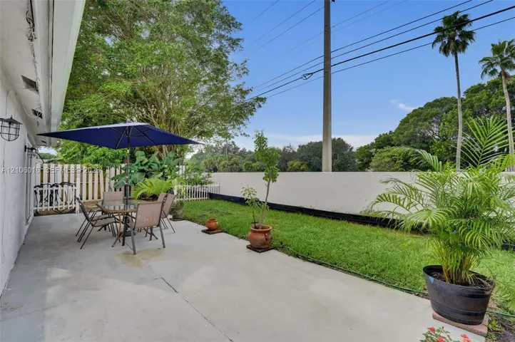 Concrete patio featuring a grass lawn, perimeter fencing, wall-mounted exterior lighting, and mature landscaping