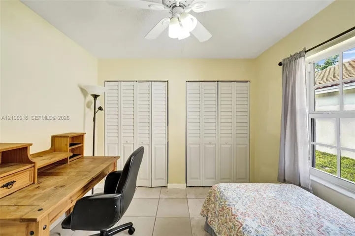 Interior room featuring light-toned ceramic tile flooring, two sets of white louvered-style bi-fold closet doors, a multi-blade ceiling fan with integrated lighting, and a single-hung window with a metal rod and drapery hardware