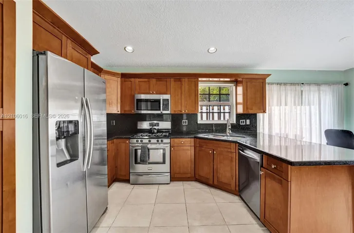 Kitchen featuring wood cabinetry, dark stone-finish countertops, stainless steel appliances, a peninsula with an integrated dishwasher, and tile flooring
