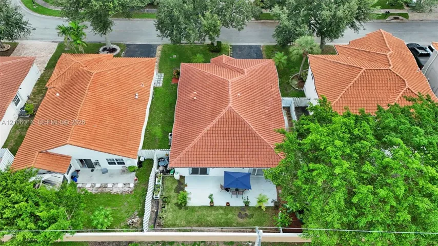 Aerial perspective of a residence featuring a terracotta-style tile roof, a paved ground-level patio, and a lawn area enclosed by a white vinyl privacy fence