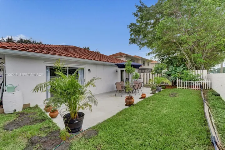 Rear exterior featuring a concrete patio, white stucco siding, a terra-cotta tile roof, and a fenced grass lawn with mature trees